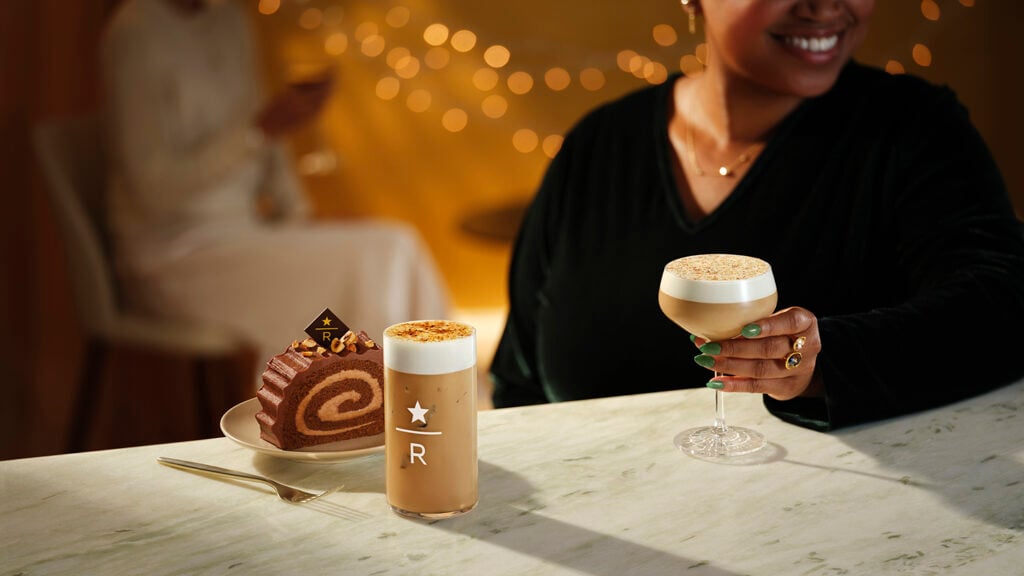 A cozy setting features a woman holding a cocktail and a glass of iced coffee, accompanied by a chocolate roll cake on a plate. Soft lighting and decorative bokeh enhance the atmosphere.