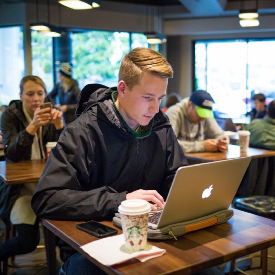 A young man in a black jacket is focused on his laptop in a coffee shop. Nearby, a woman is using her phone, and others are seated at tables. A coffee cup is on the table in front of the man.