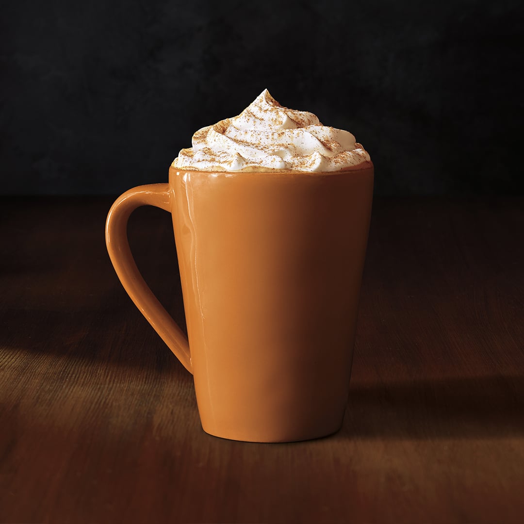 A tall, orange mug filled with creamy whipped topping, dusted with cinnamon, sits on a wooden surface against a dark background.
