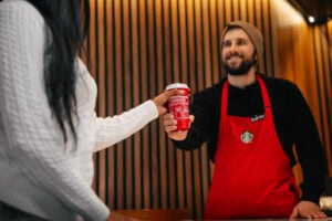 A barista in a red apron hands a red Starbucks cup to a customer wearing a white sweater. The background features wooden paneling.