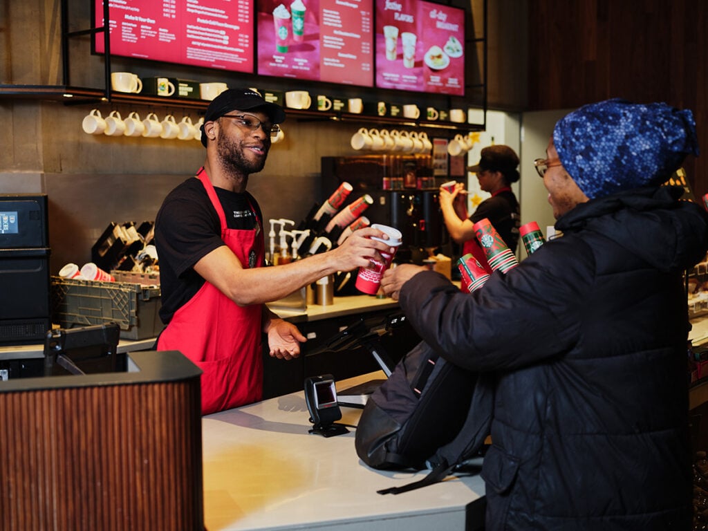 A barista in a red apron hands a coffee cup to a customer at a café counter. The customer, wearing a dark jacket and a blue patterned hat, smiles. Behind them, a menu board displays various drinks, and another staff member is preparing orders.