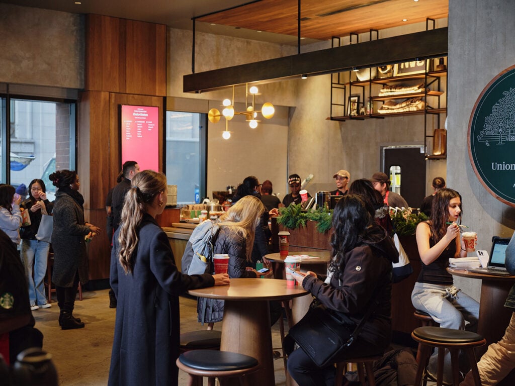 A busy café interior with patrons seated and standing. A menu board is visible, and a barista is preparing drinks. Decor includes wooden accents and greenery.