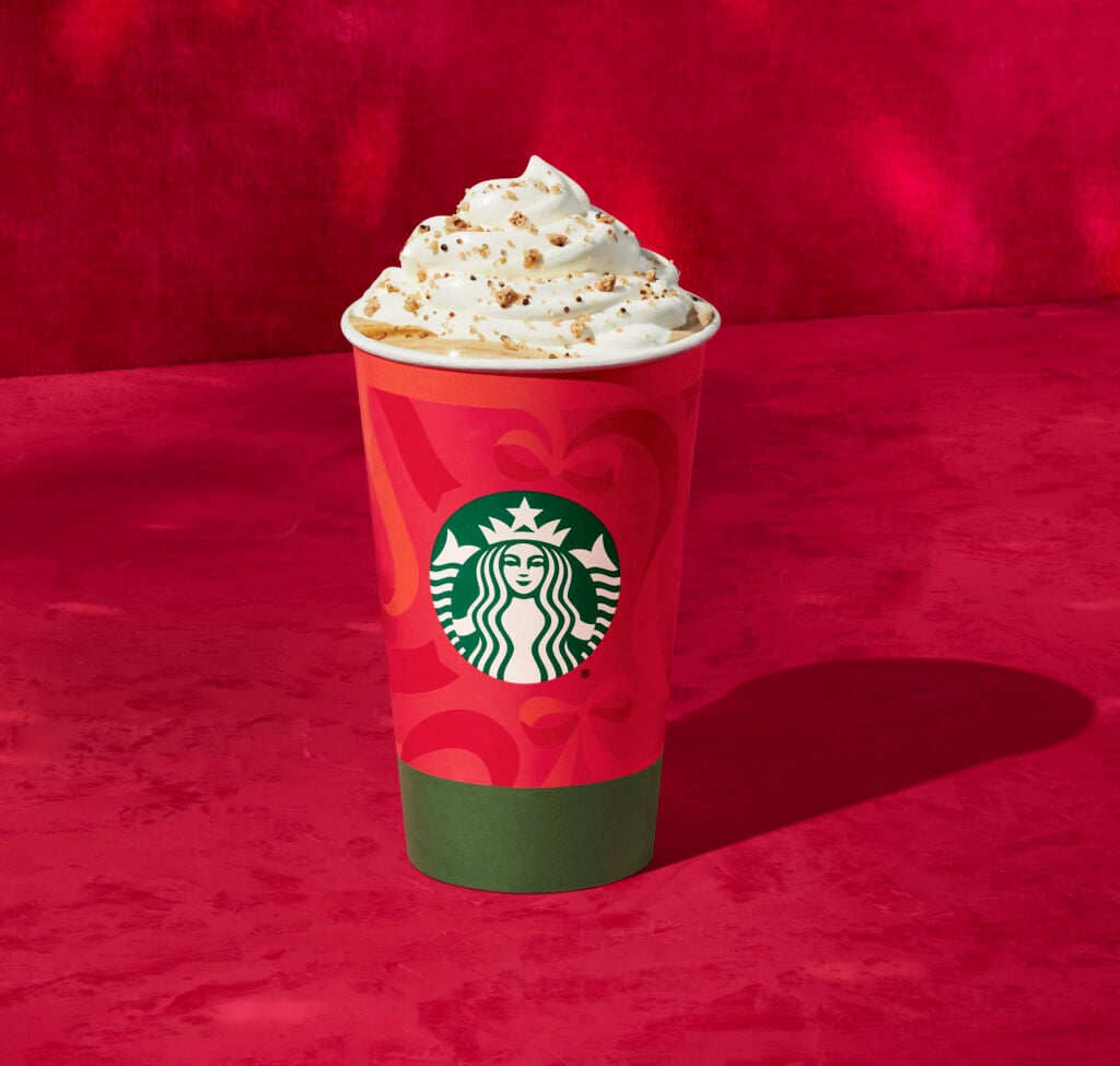 A festive red Starbucks cup topped with whipped cream and sprinkled with crumbs, set against a vibrant red background.