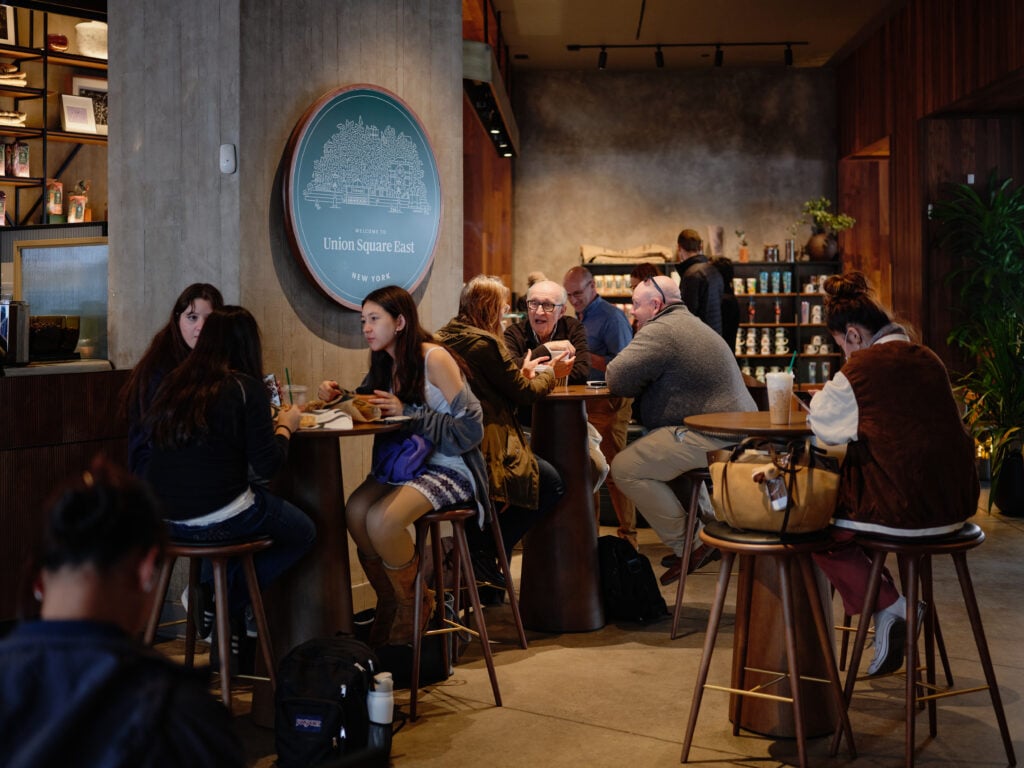 A cozy café scene featuring people seated at wooden tables, engaged in conversation and enjoying food. A sign reads "Welcome to Union Square East, New York." The interior has a warm, inviting atmosphere with a mix of casual and relaxed patrons.