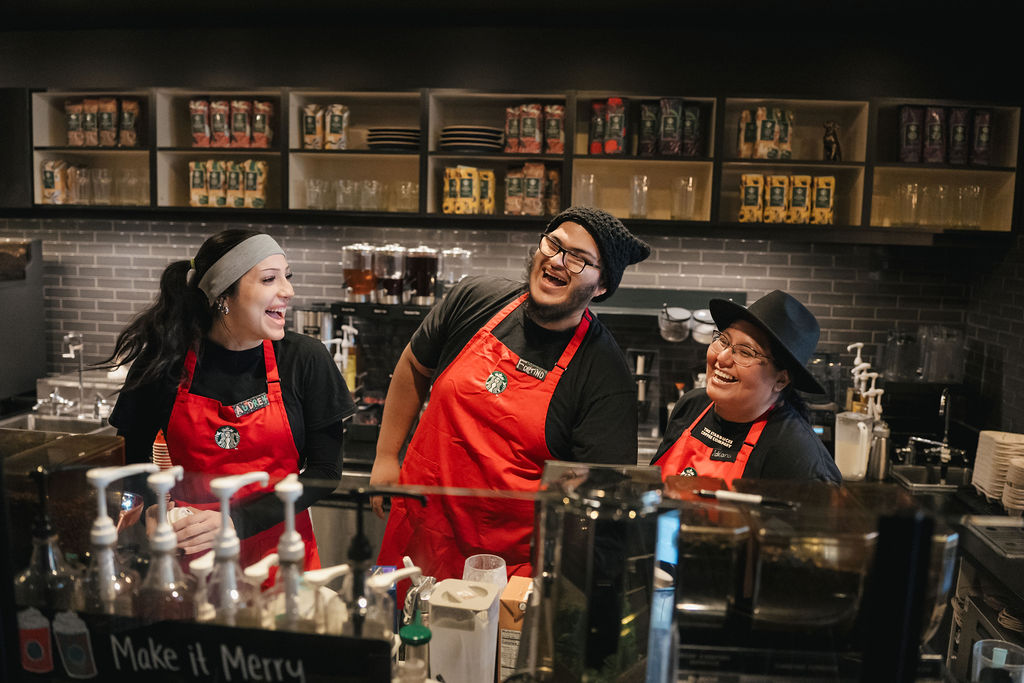 Three baristas in red aprons laugh and interact behind a coffee shop counter, with shelves of drinks and supplies in the background.