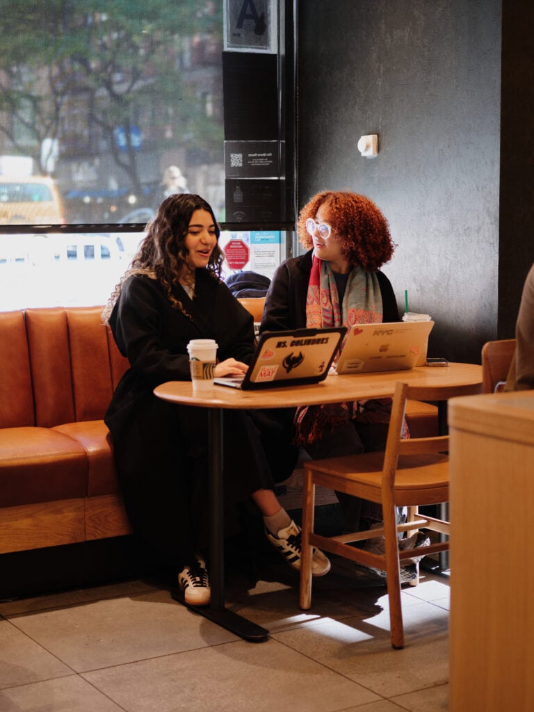 Two women are seated at a small table in a café, engaged in conversation. One has long, curly hair and is wearing a black coat, while the other has short, curly hair and is wearing glasses. Both have laptops open in front of them, and there are coffee cups on the table. The café has a cozy atmosphere with a large window showing a city view.
