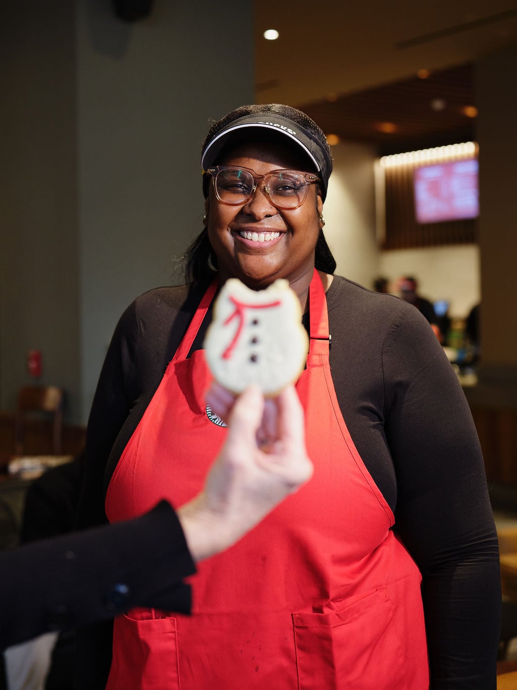 A smiling woman wearing glasses and a red apron stands in a café, holding a decorated cookie shaped like a snowman.