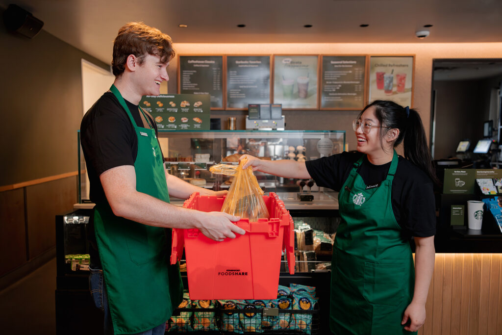 Two employees in green aprons are smiling and interacting in a café. One holds an orange food donation box while the other hands over a bag of food. The background features a display of menu items and beverages.