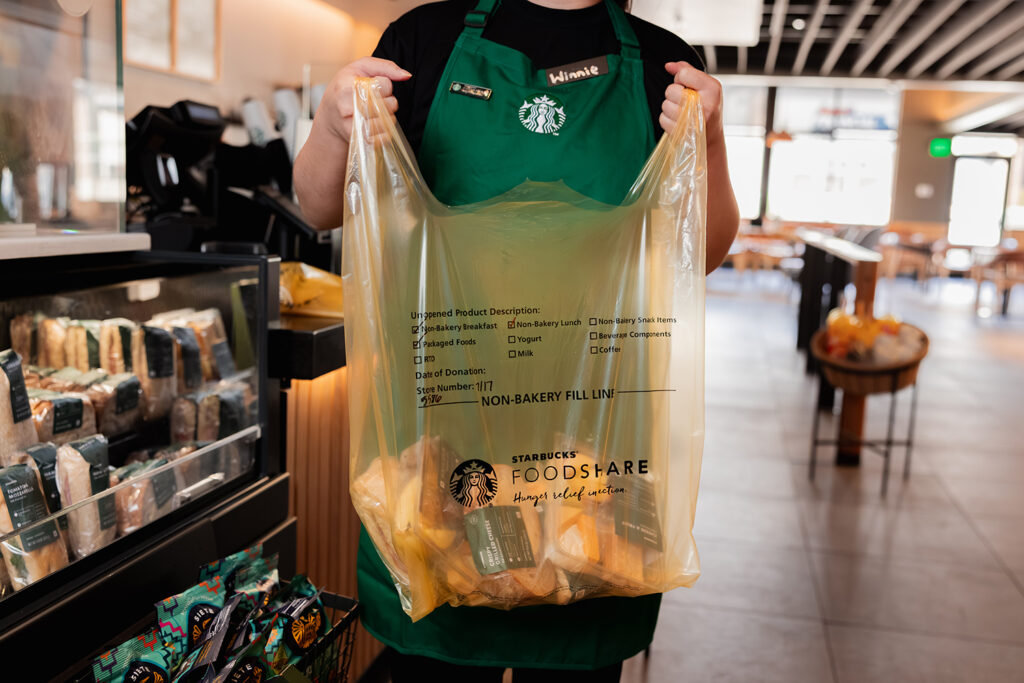 A person in a green Starbucks apron holds a large yellow bag labeled "STARBUCKS FOODSHARE," filled with food items. Behind them, a display case shows packaged snacks and drinks. The setting is a modern Starbucks interior.