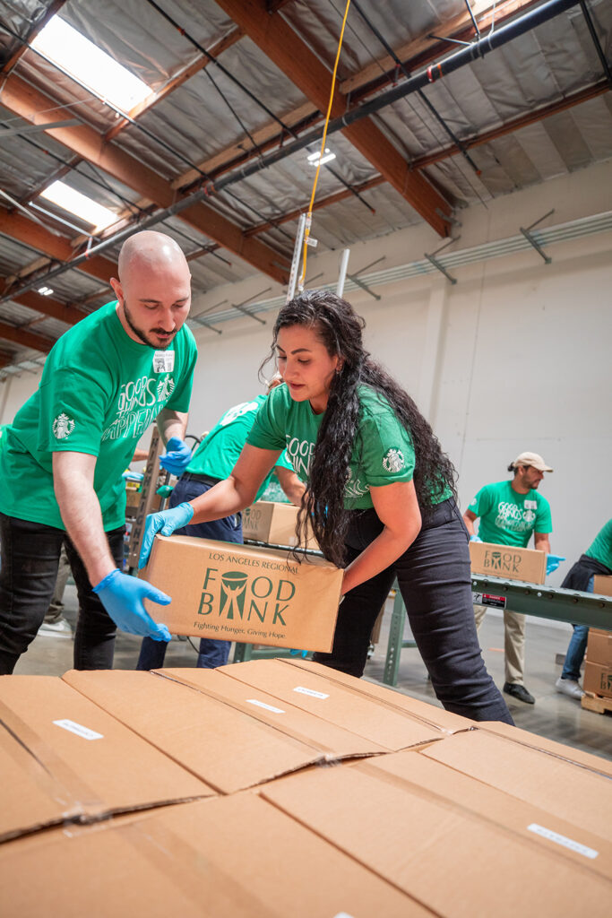 Two volunteers in green shirts are packing boxes for the Los Angeles Regional Food Bank. They are working together in a warehouse setting.