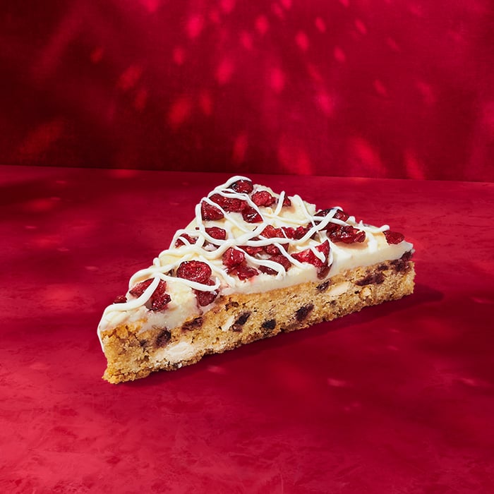 A slice of dessert featuring a cookie base topped with cream frosting, dried cranberries, and drizzled white chocolate, set against a red background.