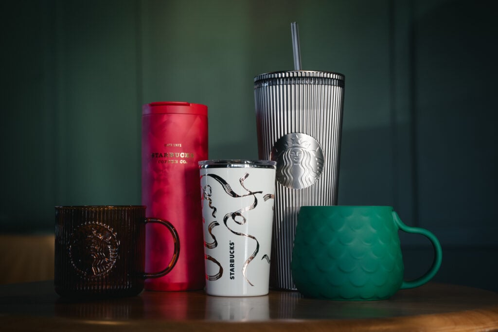 A collection of Starbucks drinkware, including a red travel tumbler, a white cup with a design, a silver striped tumbler, a brown mug, and a green textured cup.