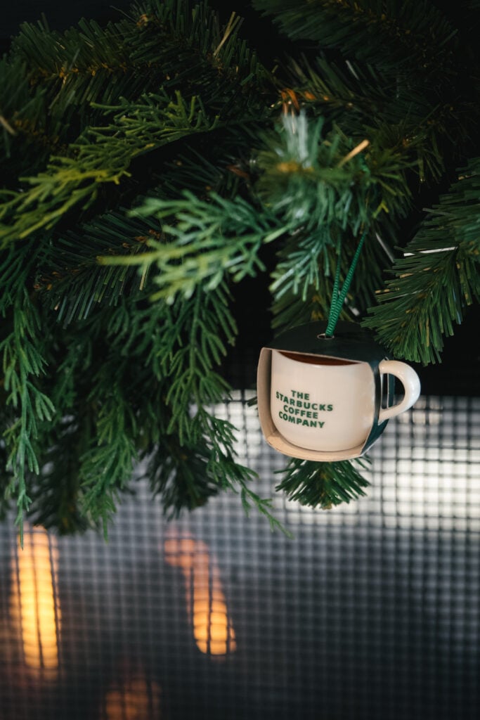 A small decorative coffee cup ornament hangs from green pine branches, featuring the text "THE STARBUCKS COFFEE COMPANY.