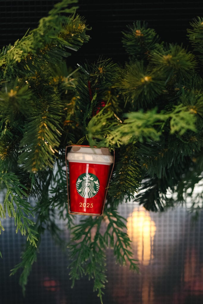 A festive red Starbucks cup ornament, labeled "2025," hangs among green pine branches, creating a holiday atmosphere.