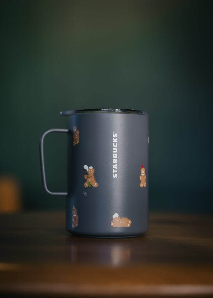 A gray Starbucks mug features playful gingerbread cookie designs and a handle, set against a blurred green background.
