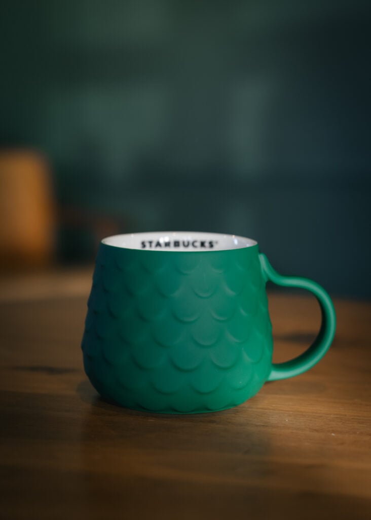 A textured green mug with a scalloped design and a white interior featuring the "STARBUCKS" logo. It rests on a wooden surface.