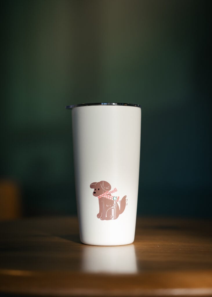 A white tumbler features a cute illustration of a dog wearing a checkered scarf, set against a blurred background.