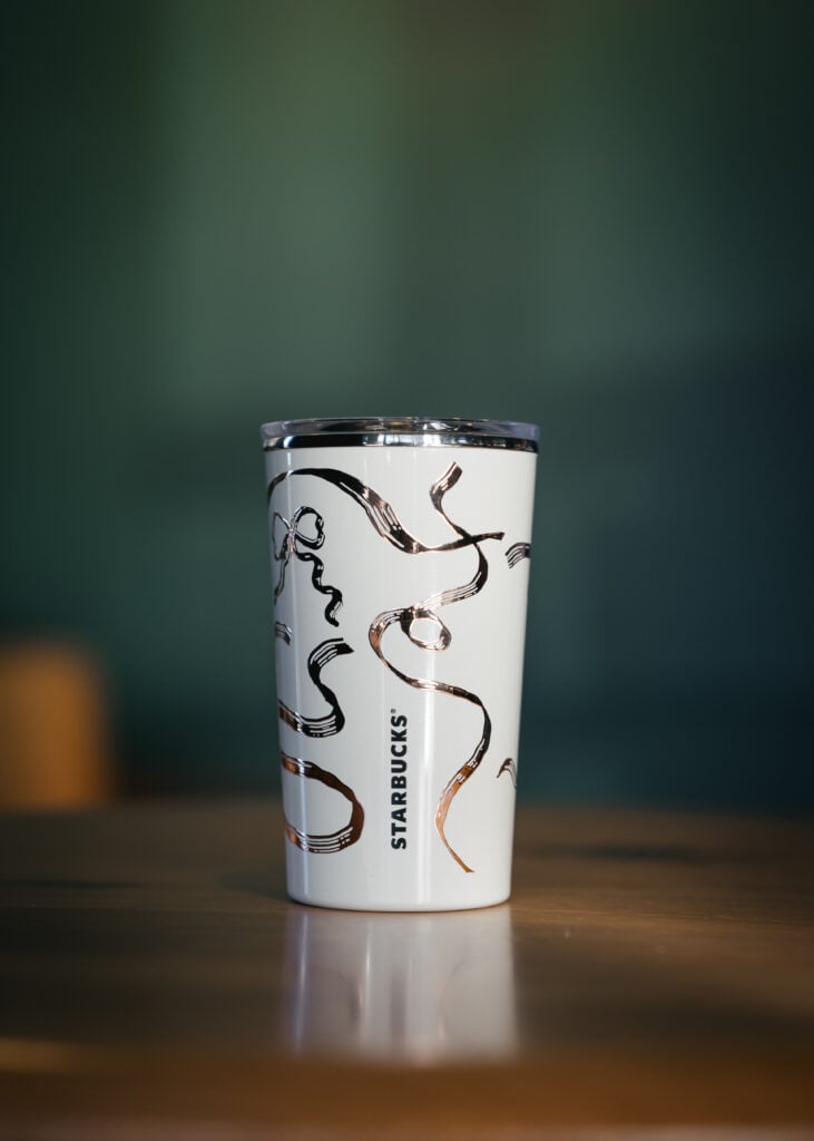 A white Starbucks tumbler with a sleek design featuring abstract, metallic rose gold lines. The background is softly blurred.