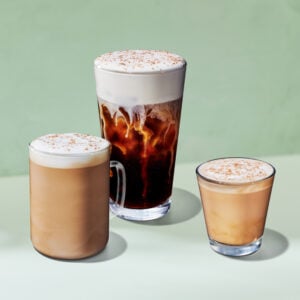 Three coffee drinks are displayed: a tall glass of iced coffee with frothy cream on top, a medium mug of creamy coffee, and a small glass of iced coffee with a light foam topping. The background is a soft green.