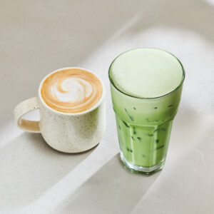 A speckled white mug holds a latte with a swirl pattern on top, next to a tall glass of iced green matcha.