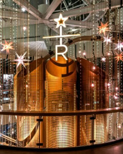 A modern interior features large, golden cylindrical structures with perforated designs. Above, a star-shaped light and hanging star decorations illuminate the space, creating a festive atmosphere.