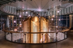 A modern interior features large, curved wooden structures with perforated designs, surrounded by hanging star-shaped lights and decorative strings of lights. The space is well-lit, showcasing a sleek railing and a contemporary aesthetic.