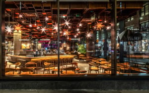 A modern restaurant interior features wooden furniture, star-shaped light fixtures, and a long bar. The ambiance is warm and inviting.