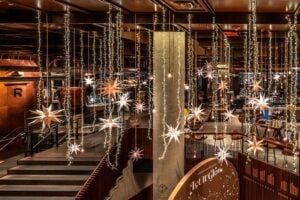 A festive interior features hanging star decorations and string lights, creating a warm, inviting atmosphere. The space includes wooden furniture and a prominent sign reading "Let It Glow.