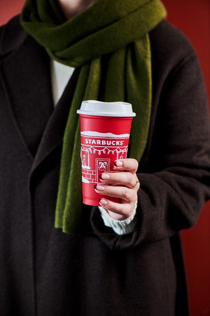 A person holds a red Starbucks cup decorated with festive designs, wearing a dark coat and a green scarf.