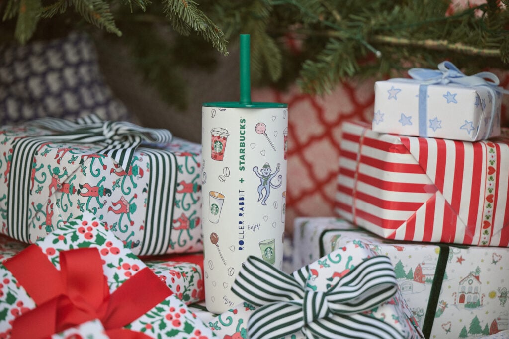 A festive Starbucks tumbler featuring playful illustrations, including drinks and a monkey, is placed among wrapped gifts with colorful patterns and ribbons.