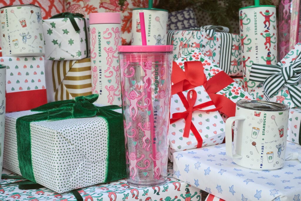 A clear tumbler with pink monkey designs and a pink straw is prominently displayed among colorful holiday gift wraps and boxes.