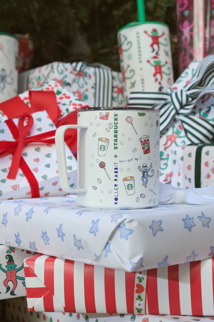 A white mug featuring playful illustrations related to coffee and treats, with "ROLLER RABBIT + STARBUCKS" printed on it. It's placed among colorful, festively wrapped gifts.