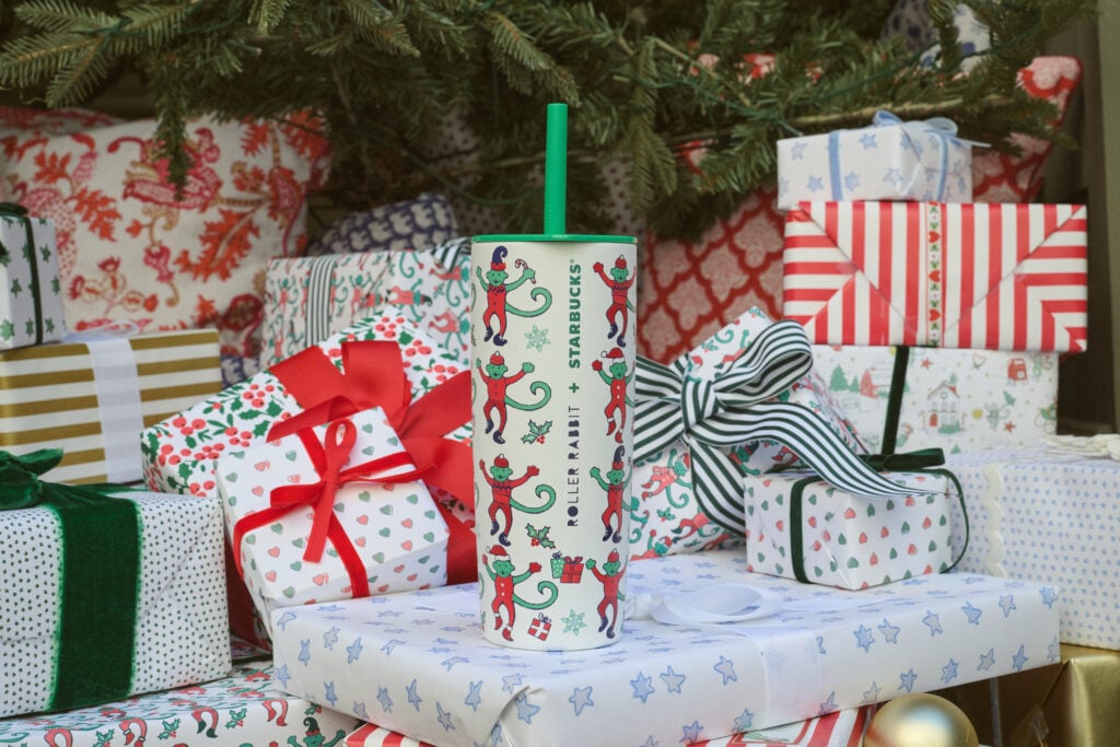 A festive Starbucks tumbler featuring playful elf designs is set against a backdrop of colorful wrapped gifts and holiday decorations.