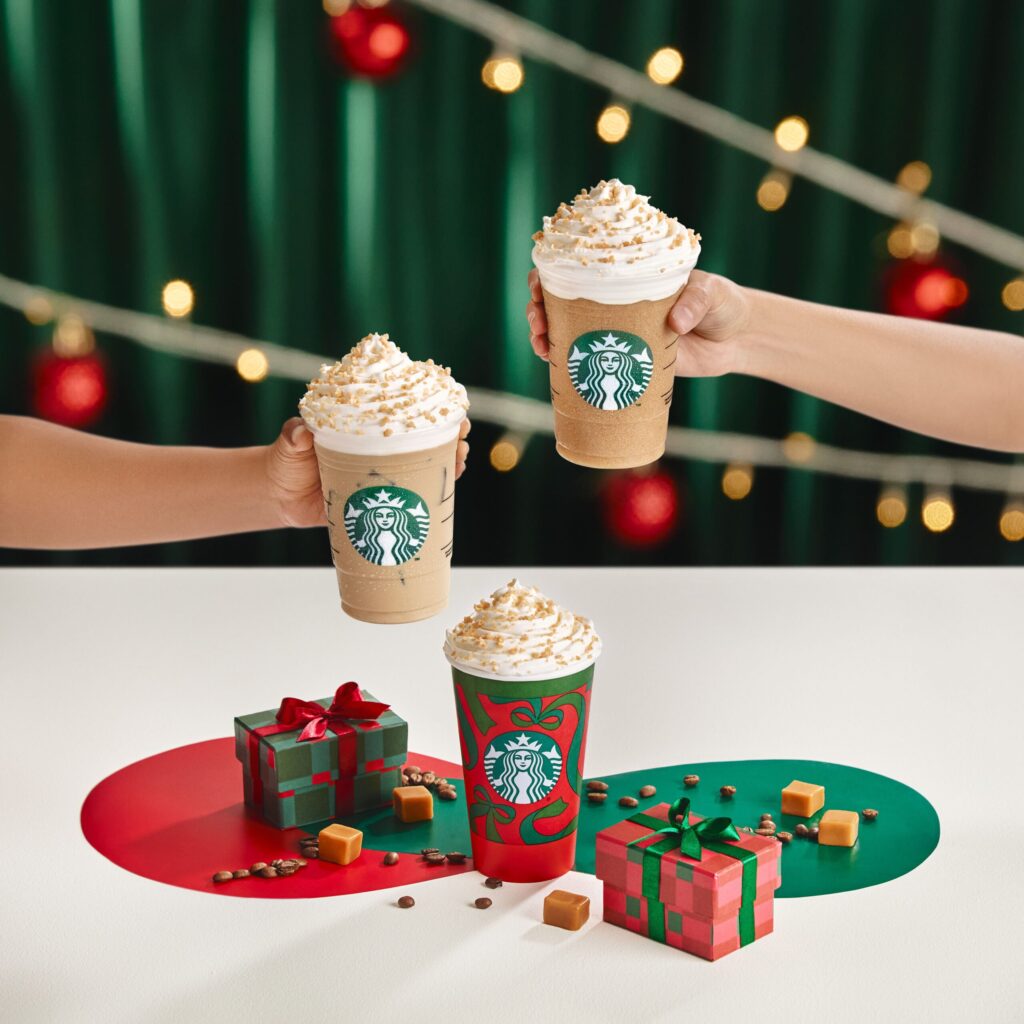 Three festive Starbucks drinks topped with whipped cream and caramel are held up against a backdrop of holiday decorations. Colorful gift boxes and caramel candies are arranged on a table, enhancing the seasonal theme.
