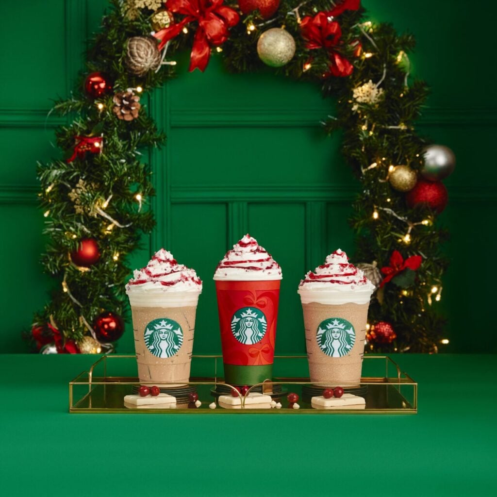 Three festive Starbucks drinks are displayed on a gold tray against a green backdrop. The drinks feature whipped cream and red toppings, with one in a red cup and two in clear cups. A decorated holiday wreath with ornaments and lights is in the background.