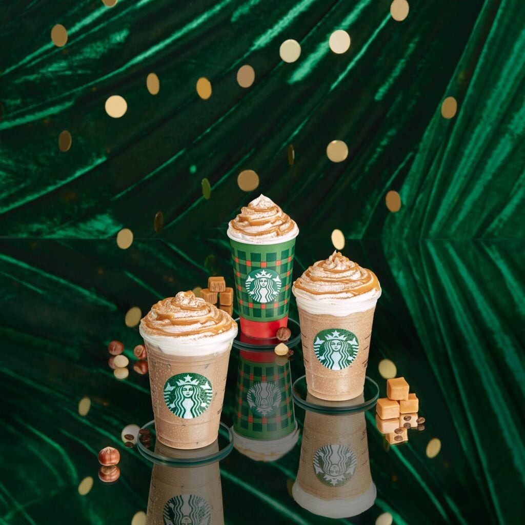 Three Starbucks drinks are displayed against a green velvet backdrop with gold confetti. Each drink features whipped cream and caramel drizzle, with one in a festive green and red cup. Surrounding the cups are caramel pieces and coffee beans.