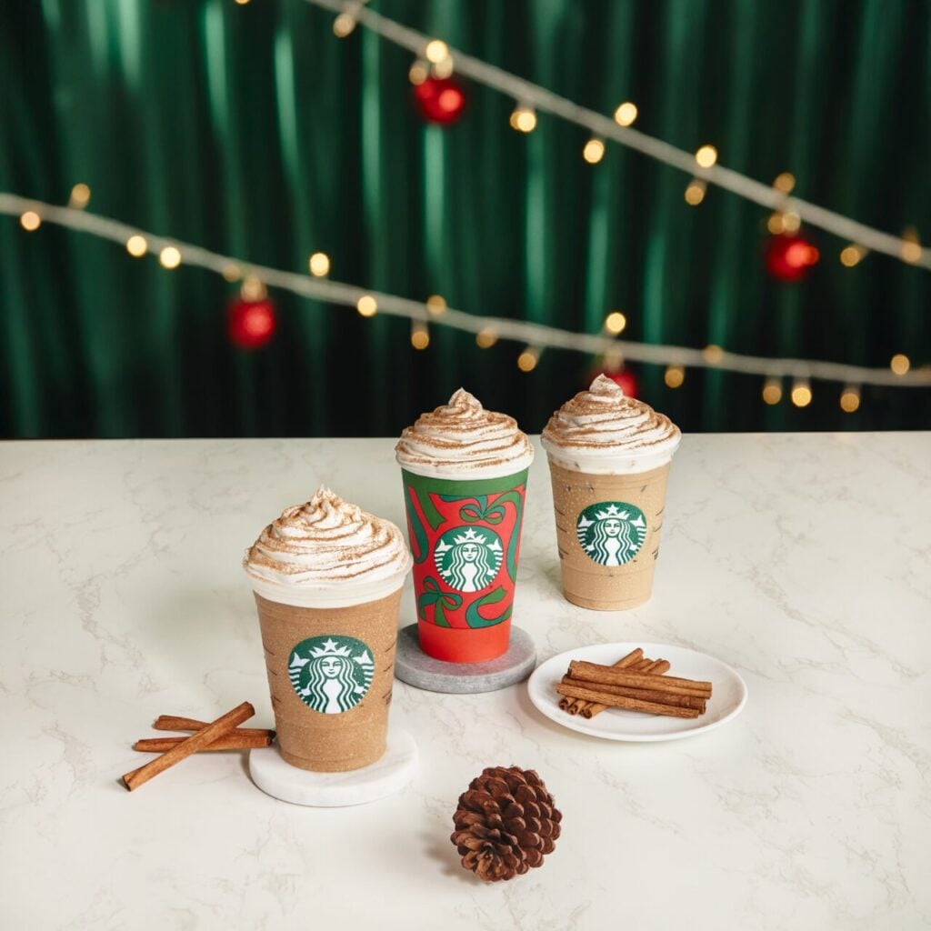 Three festive Starbucks cups topped with whipped cream and cinnamon sit on a marble surface, accompanied by cinnamon sticks and a pinecone. A green backdrop with string lights and red ornaments adds to the holiday atmosphere.
