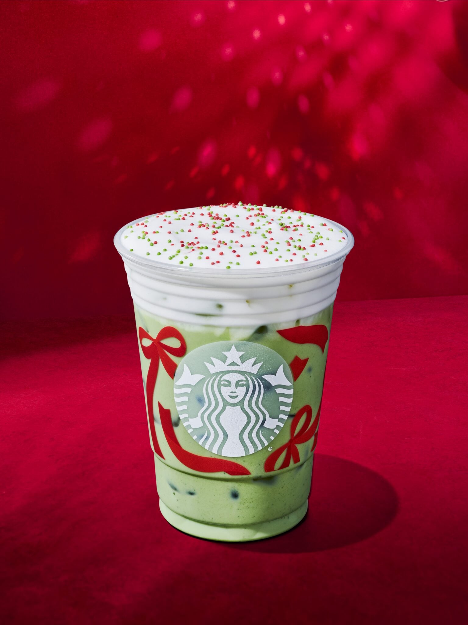 A festive Starbucks cup filled with a green beverage, topped with whipped cream and red and green sprinkles, set against a red background.