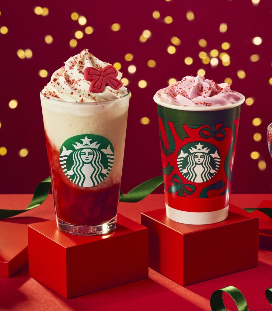 Two festive Starbucks drinks are displayed on red pedestals. The left drink features whipped cream topped with a red bow and strawberry sauce. The right drink is in a decorated cup with pink whipped cream and red sprinkles. A blurred background of golden lights enhances the holiday theme.