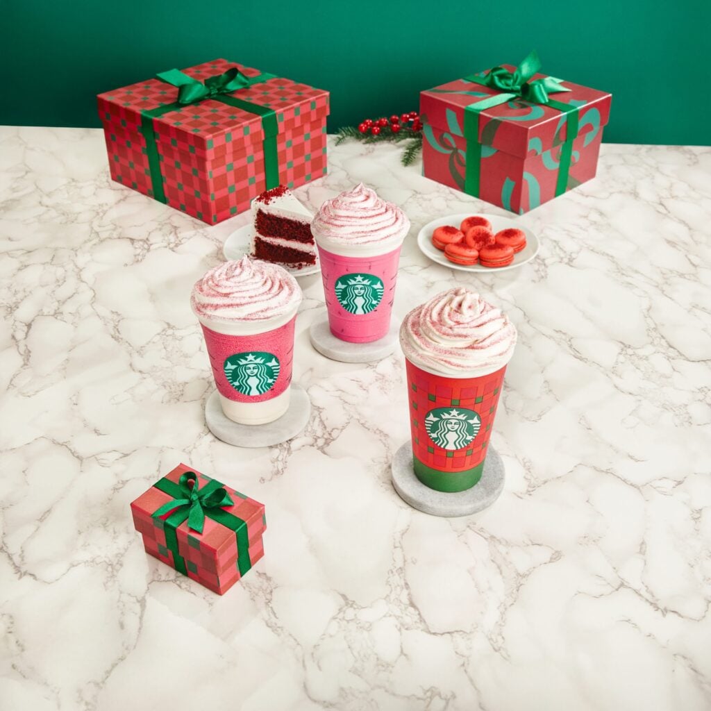 Three festive Starbucks drinks with whipped cream sit on a marble table, accompanied by a small gift box, red velvet cake, and macarons.