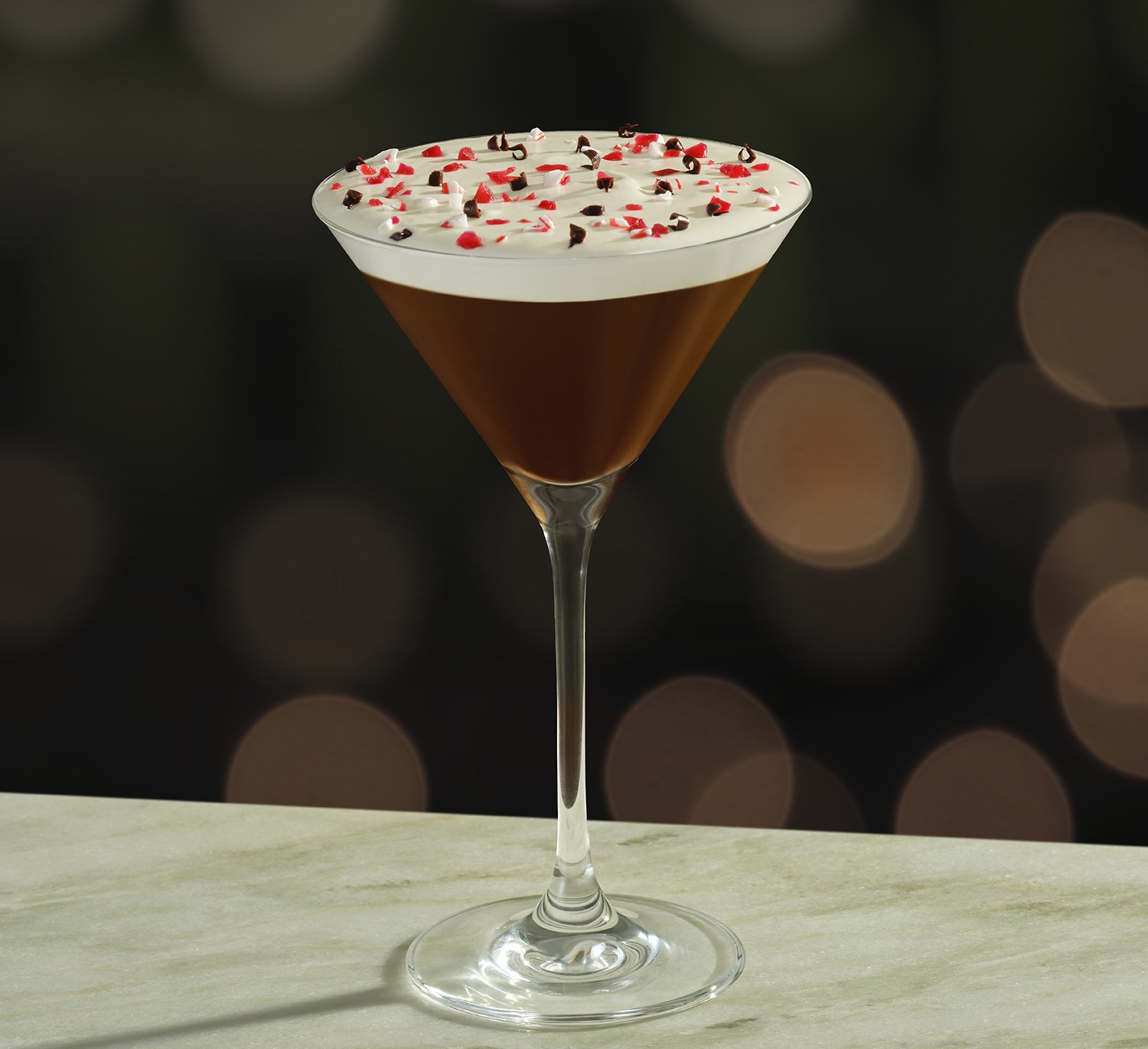 A martini glass filled with a dark brown beverage, topped with whipped cream, red and white sprinkles, and chocolate shavings.