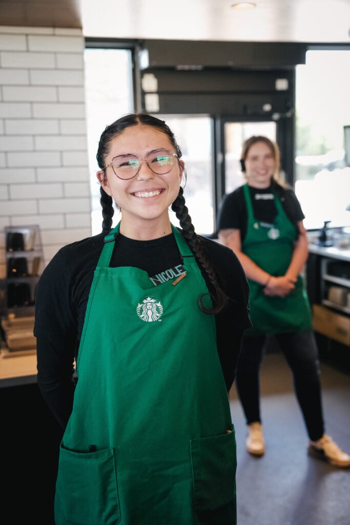 A smiling barista with braided hair wears a green Starbucks apron and glasses. Another staff member stands in the background.