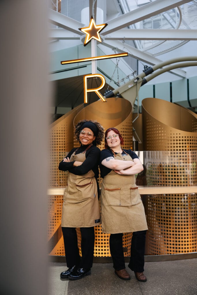 Two individuals in brown aprons stand confidently with arms crossed, smiling. Behind them is a decorative backdrop with a glowing star and the letter "R." The setting features modern design elements.