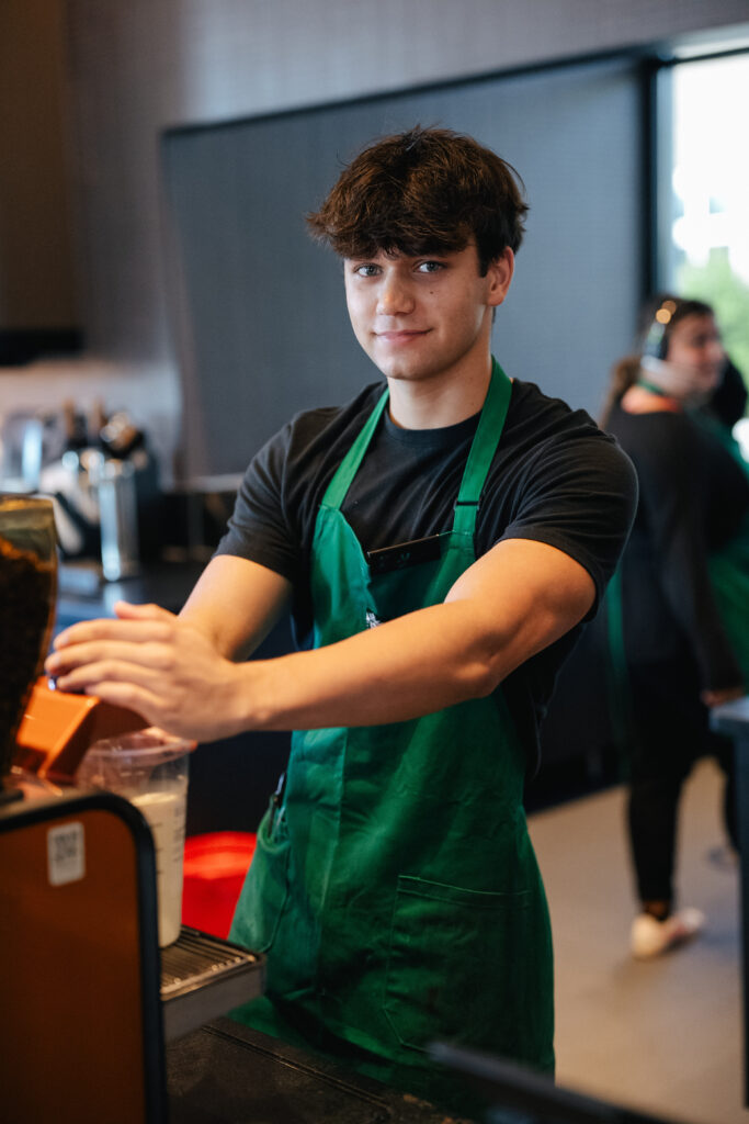 A young barista in a green apron stands at a coffee counter, preparing a drink. He has short, tousled hair and is smiling.