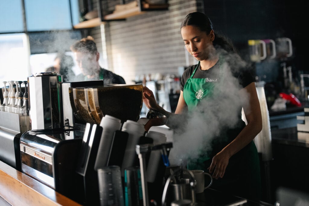 A barista in a green apron prepares coffee at a café, with steam rising from the equipment. Another worker is visible in the background.
