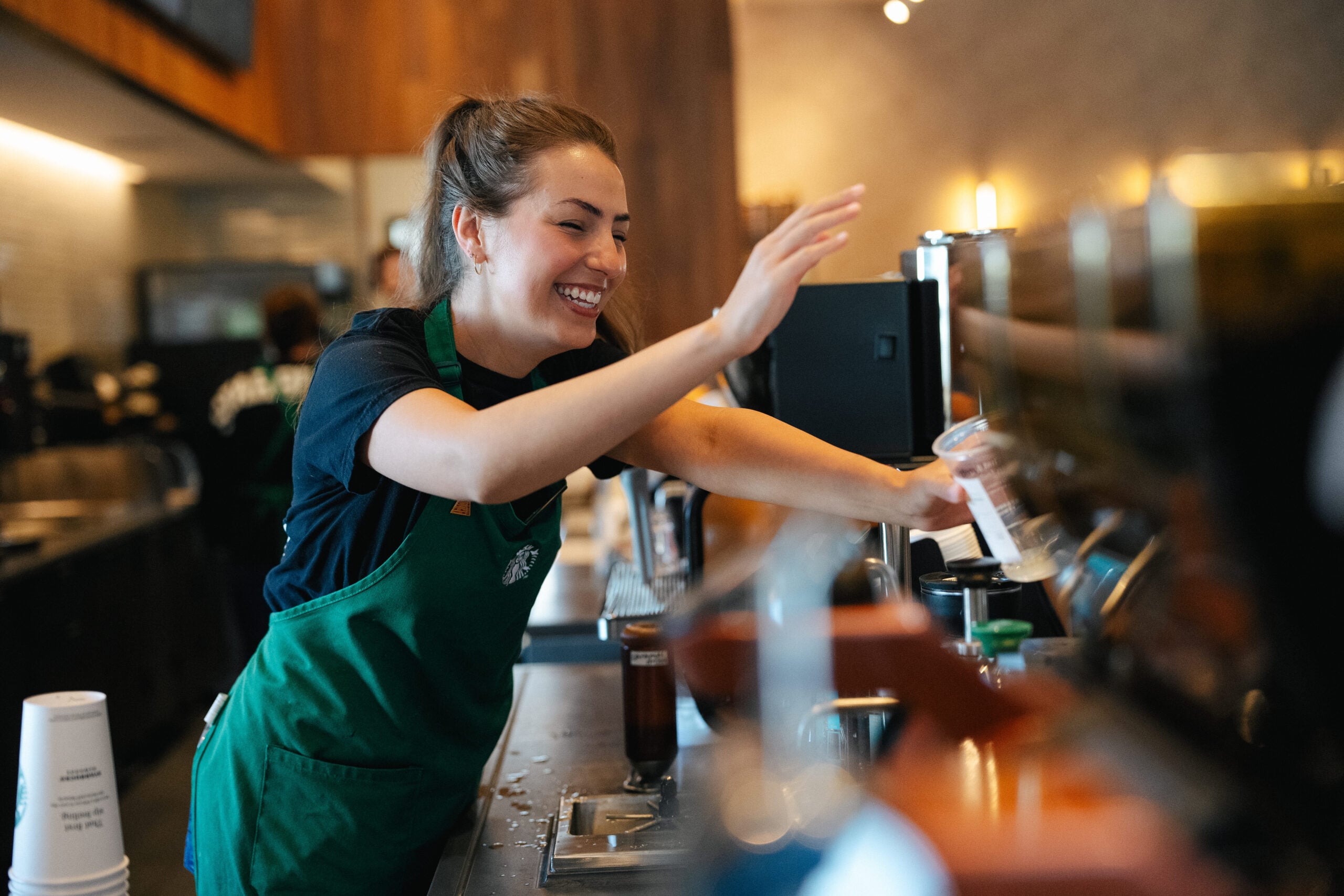 A barista in a green apron smiles while serving coffee at a café, with a lively atmosphere and various coffee equipment in the background.