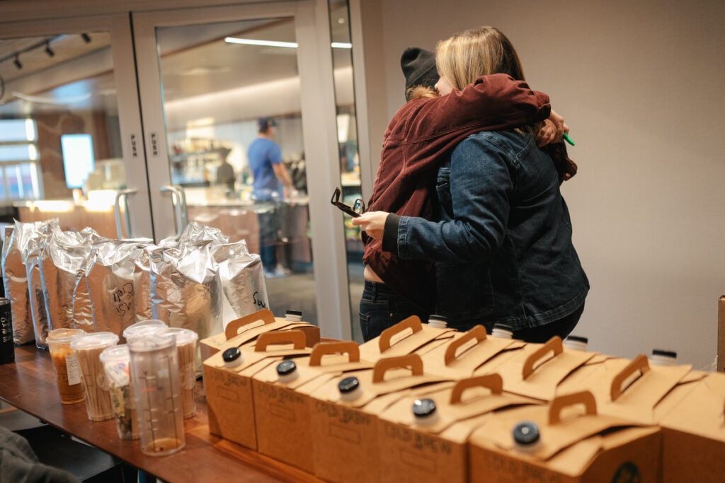 Two people are embracing in a casual setting, with a table in the foreground displaying several cartons labeled "COLD BREW" and some silver packages. In the background, a person is visible near a counter.