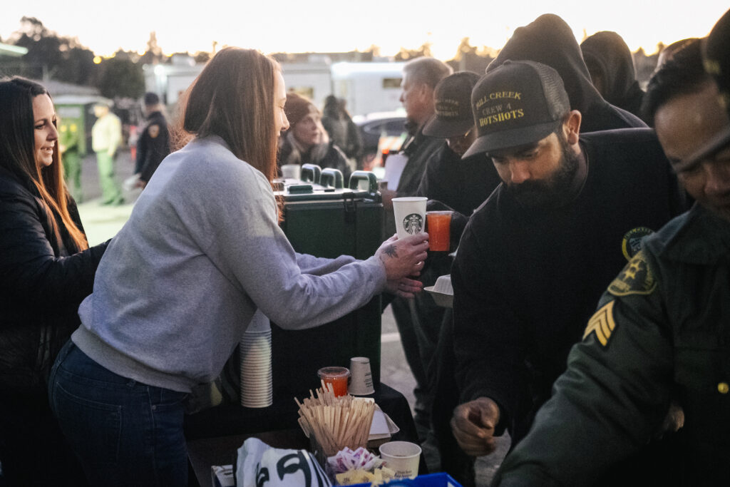 A woman hands a coffee cup to a man at a food station, with others in the background. The scene is set outdoors during twilight.