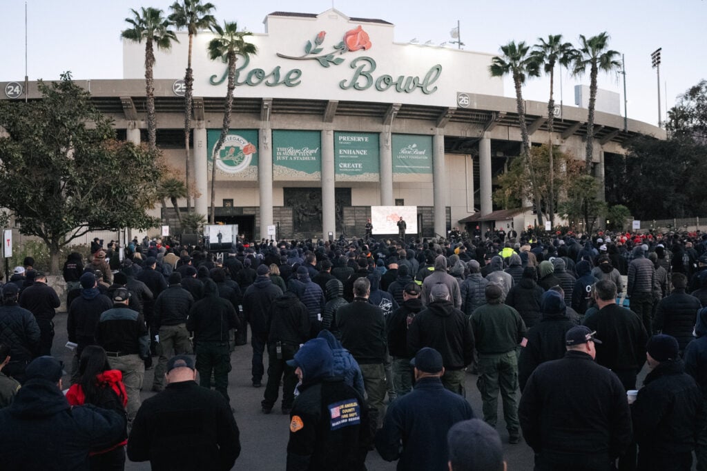 A large crowd gathers in front of the Rose Bowl, with many people wearing dark jackets and hooded sweatshirts. Palm trees are visible nearby, and a stage is set up with speakers addressing the audience.