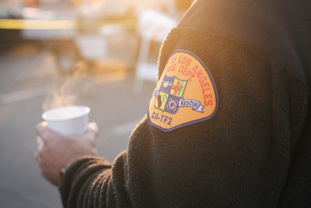A close-up of a person's arm wearing a black fleece jacket with an embroidered patch of the Los Angeles Fire Department. The patch features various symbols and the text "CA-TF2" and "RESCUE." The person is holding a steaming cup.